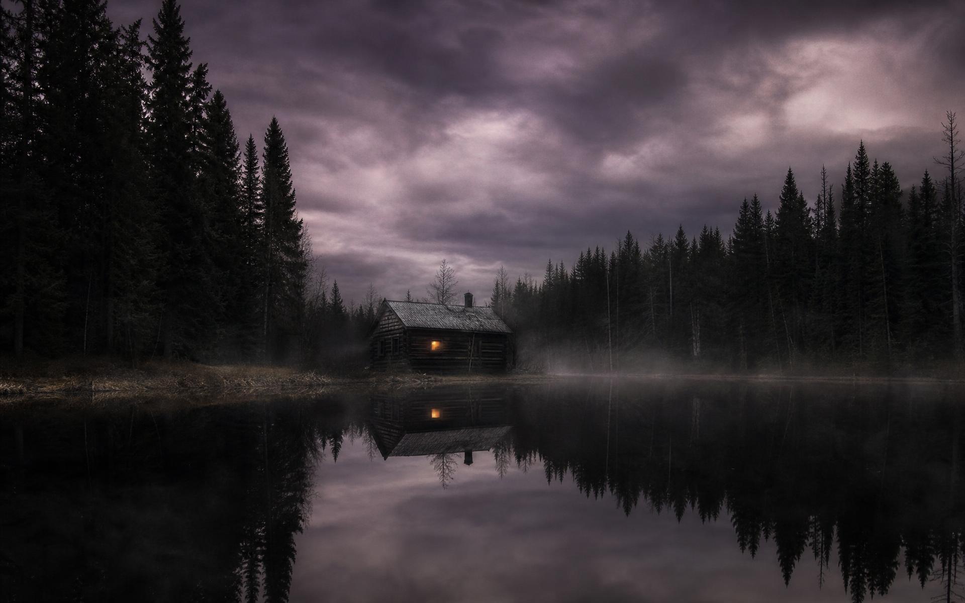 An abandoned log cabin at the edge of a black lake, pine forest closing in, one amber window glowing beneath a bruised sky